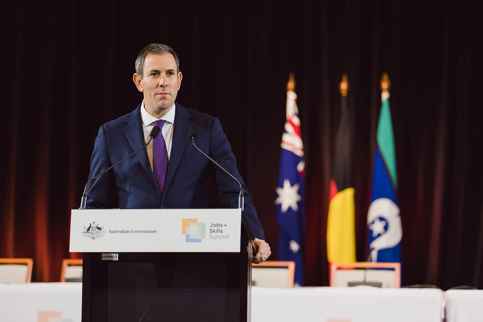 Australian Treasurer Jim Chalmers speaking at a government lectern, with the Australian, Aboriginal and Torres Strait Islander flags behind him.