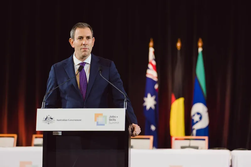 Australian Treasurer Jim Chalmers speaking at a government lectern, with the Australian, Aboriginal and Torres Strait Islander flags behind him.